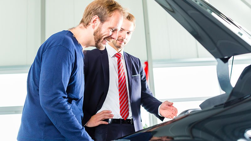 To menn står ved et åpent bagasjerom på en bil. Den ene mannen smiler og ser ned i bagasjerommet, den andre er kledd i dress med slips, og er fokusert på den første mannen.