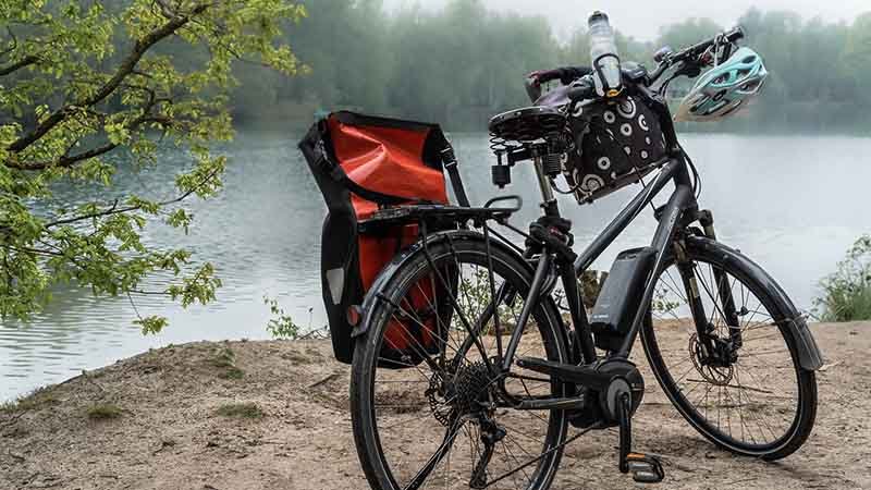 Elsykkel med sidevesker, hjelm og drikkeflaske står parkert ved vannkanten med trær i bakgrunnen.