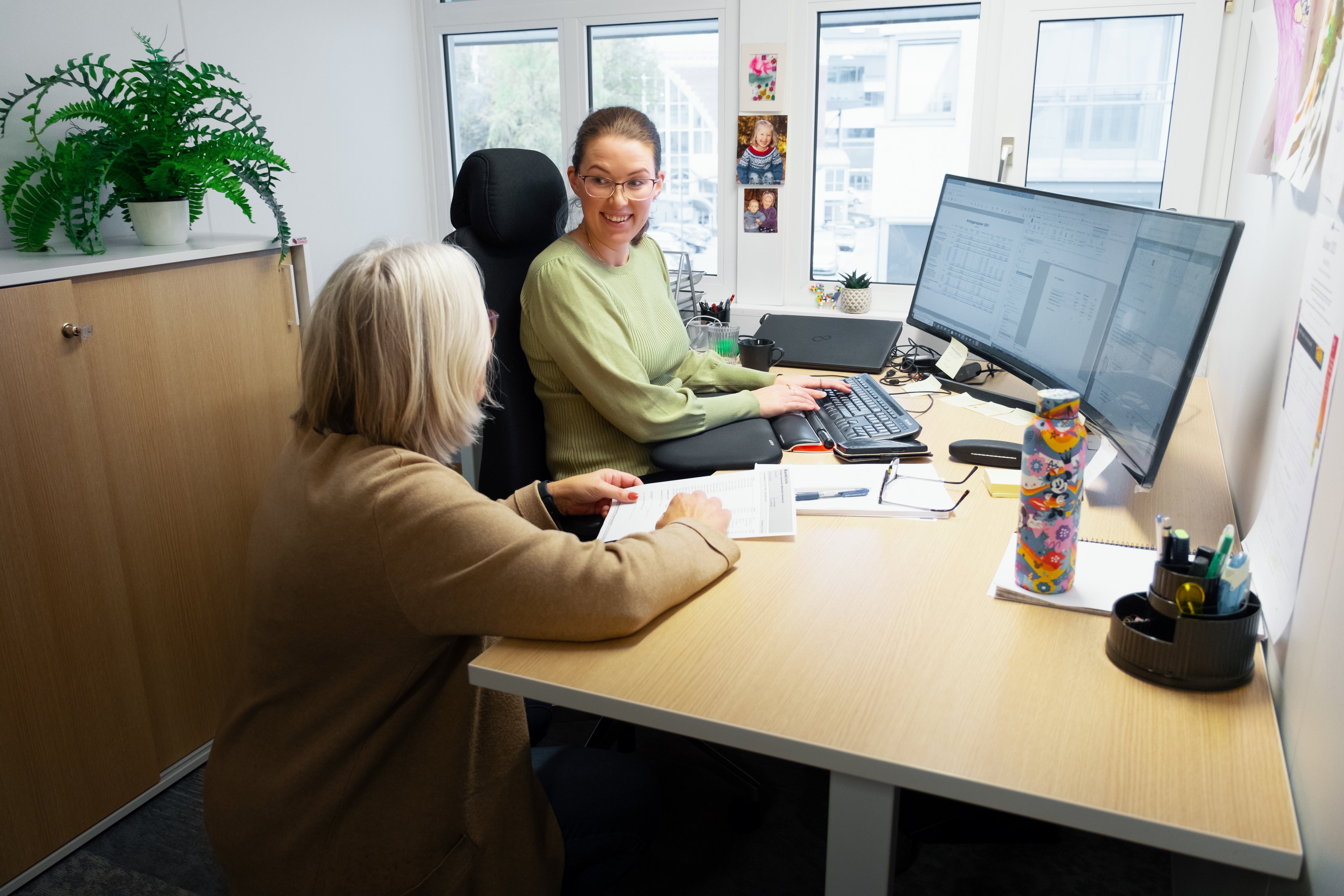 To kvinner samarbeider ved en kontorpult foran en dataskjerm.
