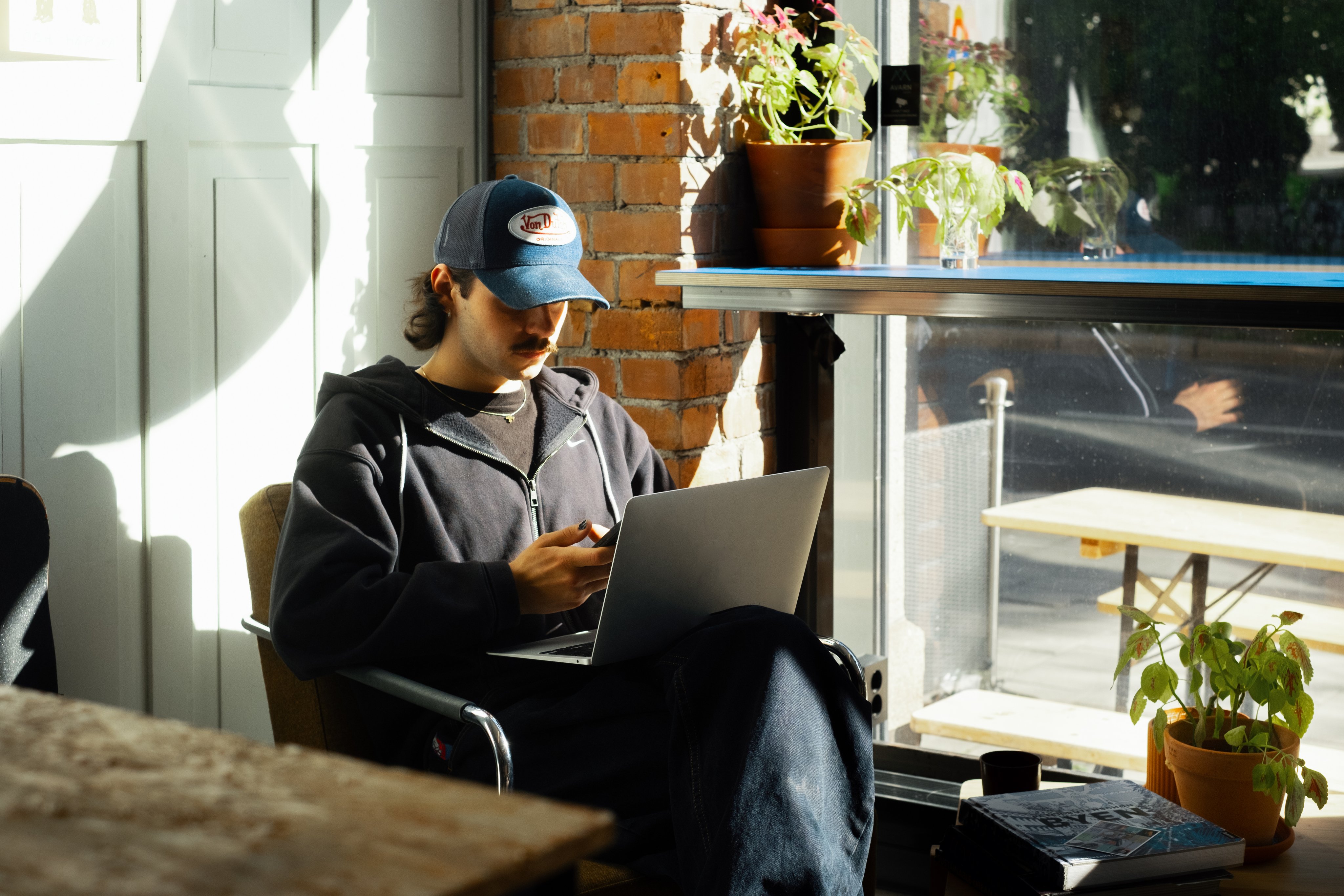 En ung mann sitter i en solfylt kafé ved et vindu og arbeider på en bærbar PC mens han holder en mobiltelefon.
