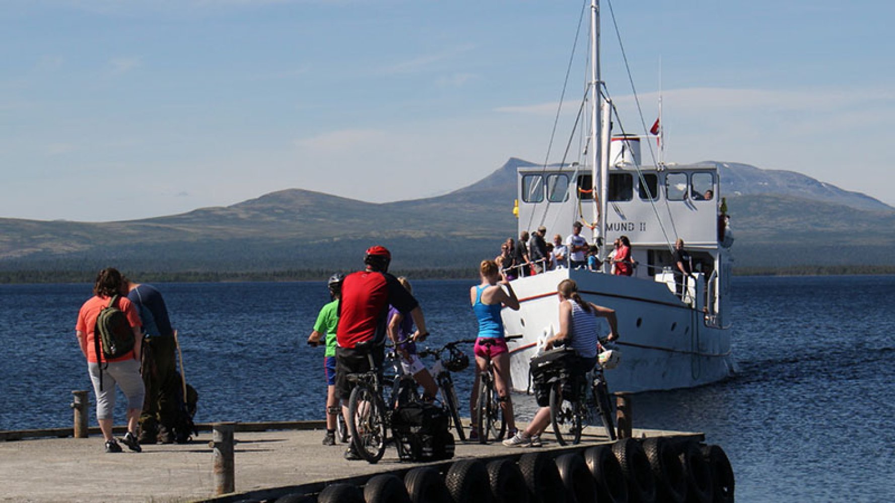 Syklister og fotgjengere står på en brygge mens en passasjerbåt nærmer seg, med fjell og blått vann i bakgrunnen.