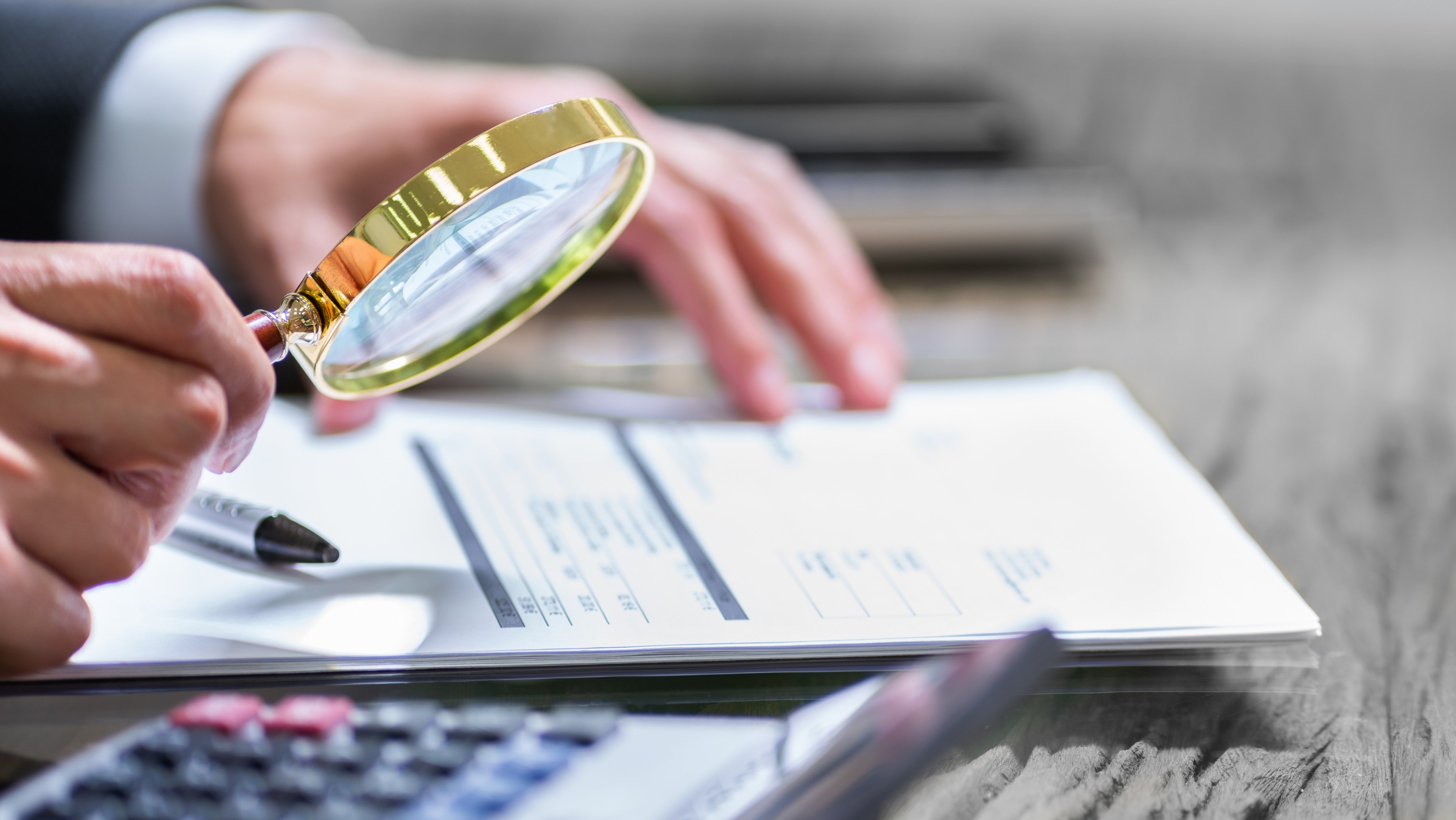 En hånd holder et forstørrelsesglass over noen dokumenter på et bord, ved siden av ligger en kalkulator