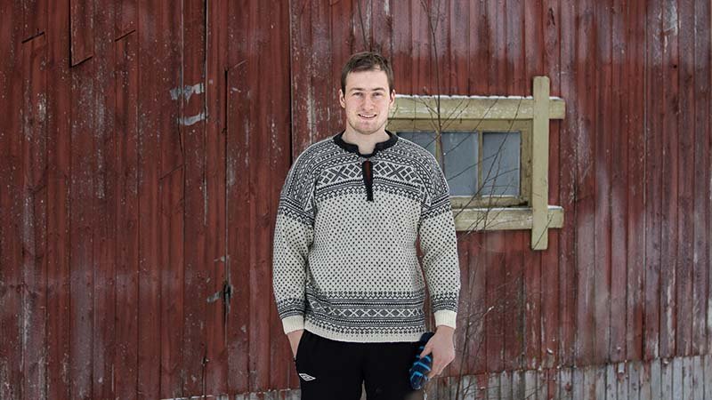 Mann i strikket genser som står foran en rød låve og smiler.