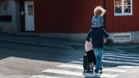 En mann bærer et barn og triller en koffert på vei over et fotgjengerfelt.