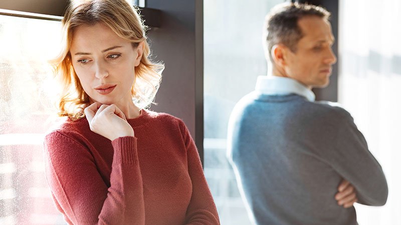 Et par står innendørs med ryggen delvis vendt mot hverandre, ved et stort vindu.