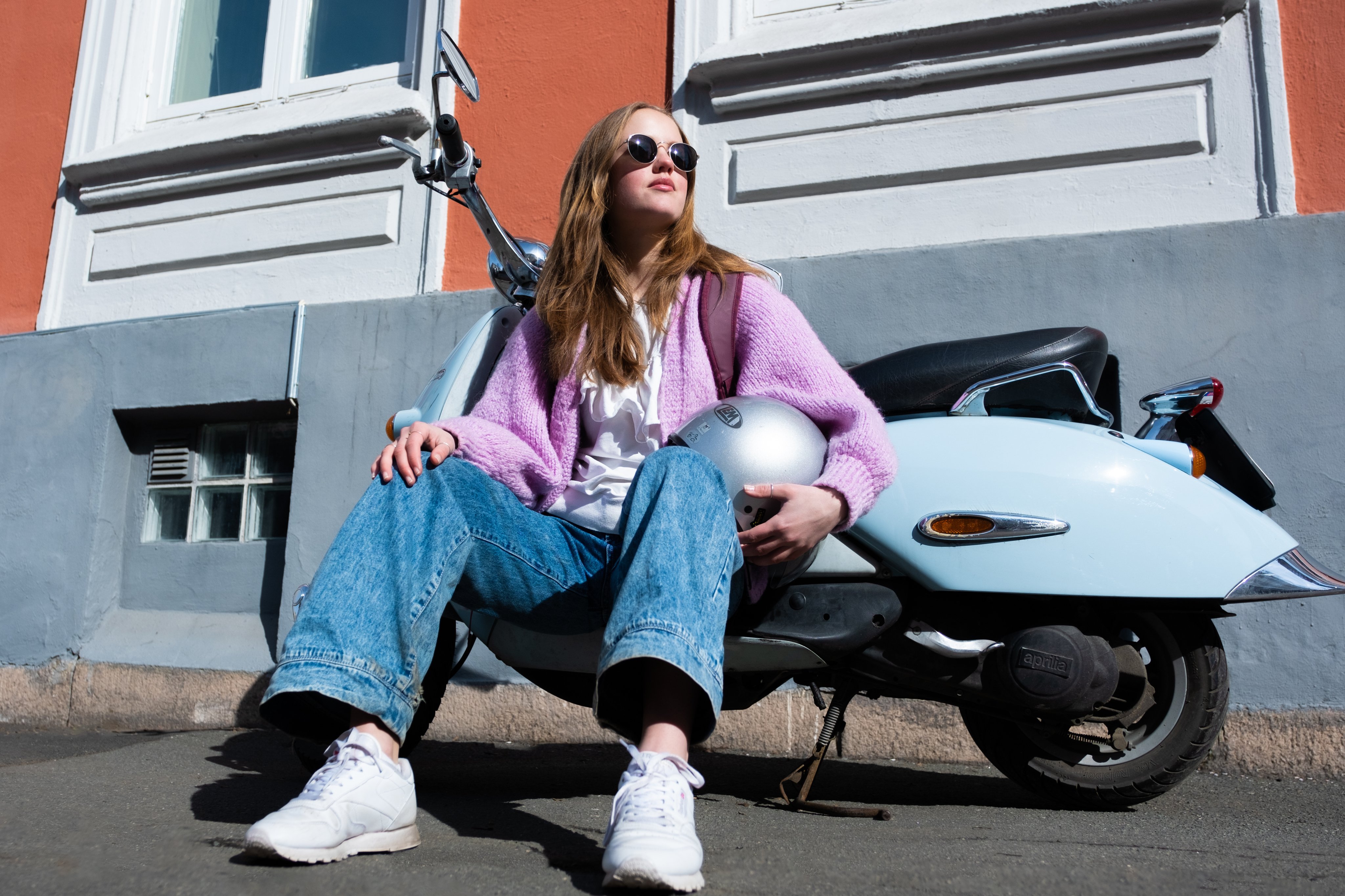 Ung kvinne sitter på bakken foran en scooter og holder en hjelm.