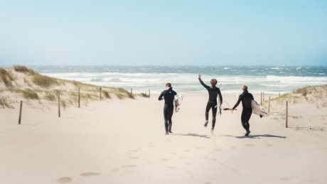 Tre surfere med surfebrett på vei mot havet langs en sandstrand.