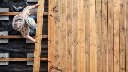 En mann legger terrassebord på en ramme av bjelker, med verktøy og skruer synlig.