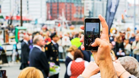 En hånd holder en mobiltelefon og tar bilde av en folkemengde. 