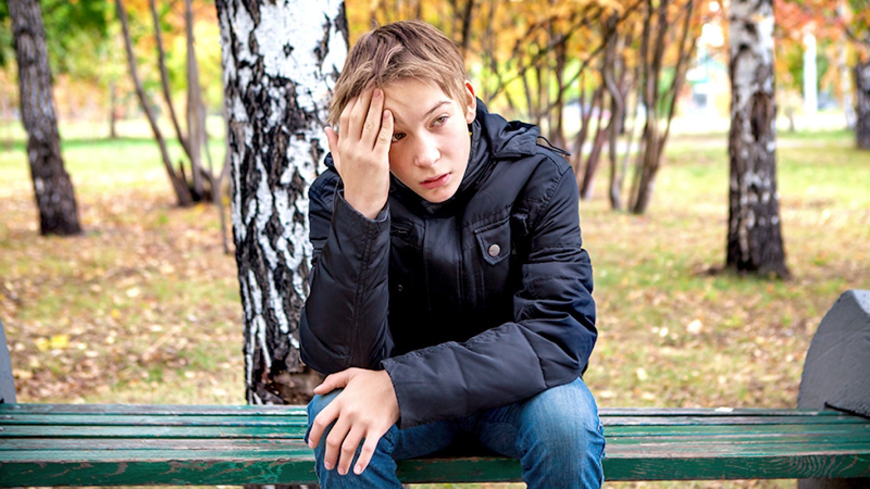 En gutt sitter på en  benk i en park med høstfargede trær i bakgrunnen. Han ser trist ut.