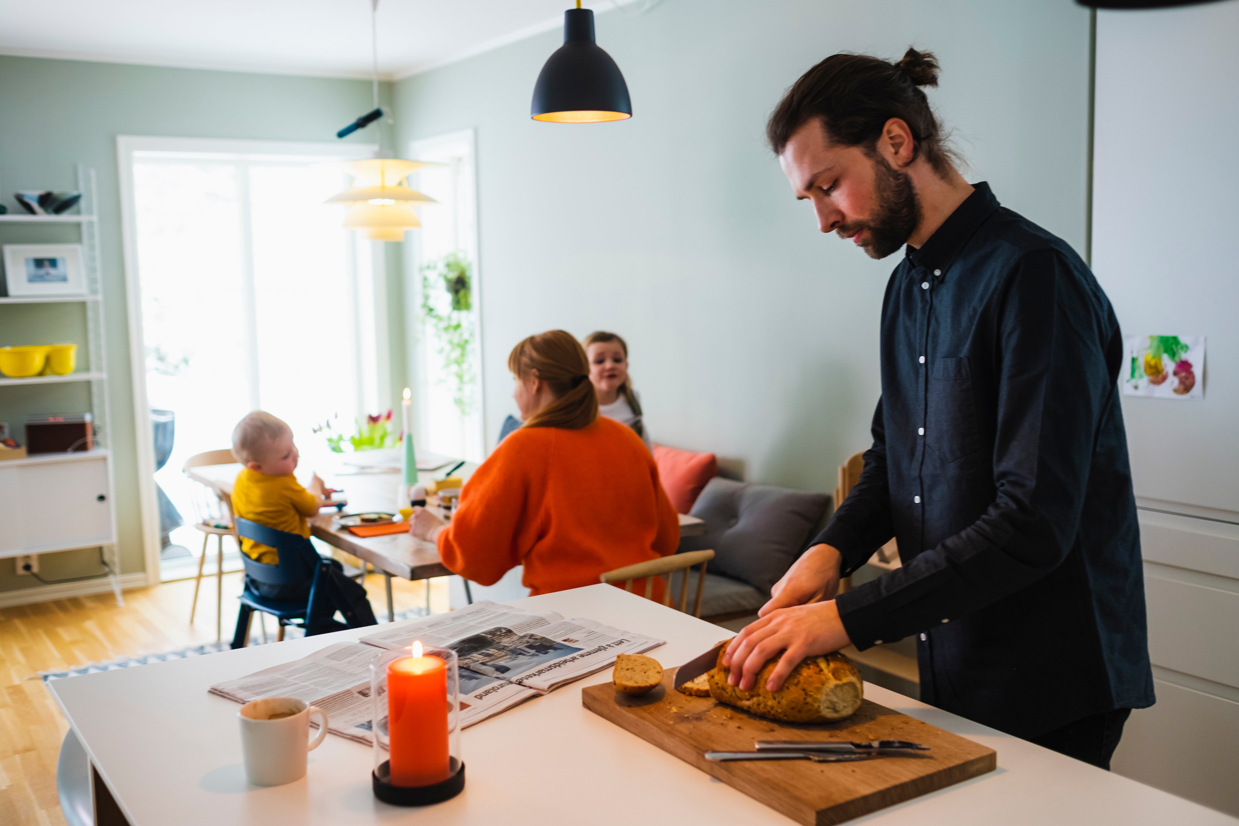 En mann står på et kjøkken og kutter brødskiver, med kone og barn på et spisebord i bakgrunnen.