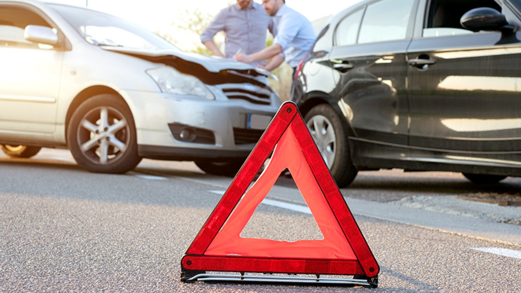 Nærbilde av en varseltrekant med to biler i bakgrunnen som har kollidert. To menn står og skriver skademelding på panseret