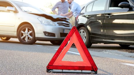 Nærbilde av en varseltrekant med to biler i bakgrunnen som har kollidert. To menn står og skriver skademelding på panseret