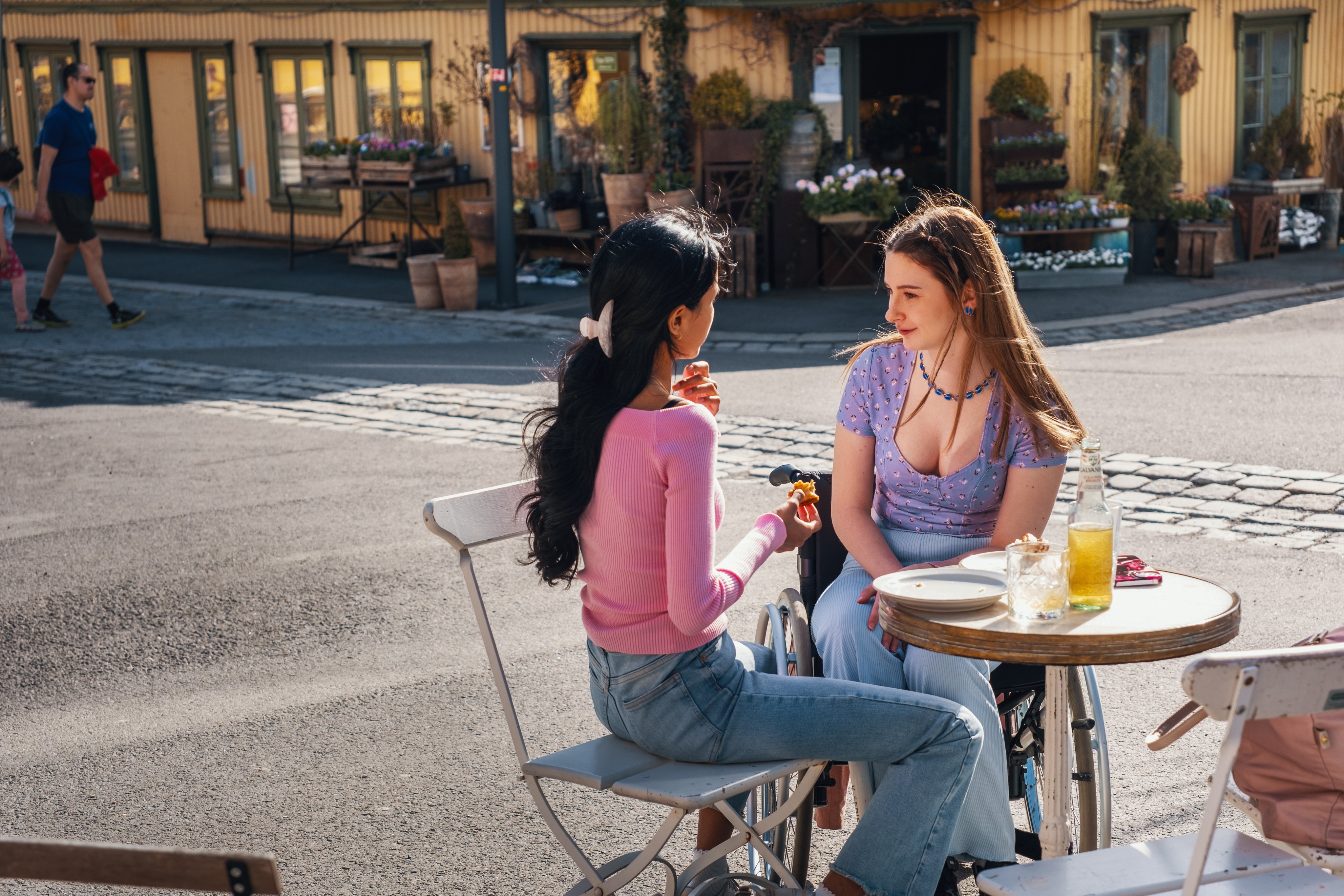 To unge kvinner sitter ved et kafébord utendørs og spiser, med drikke og mat på bordet.