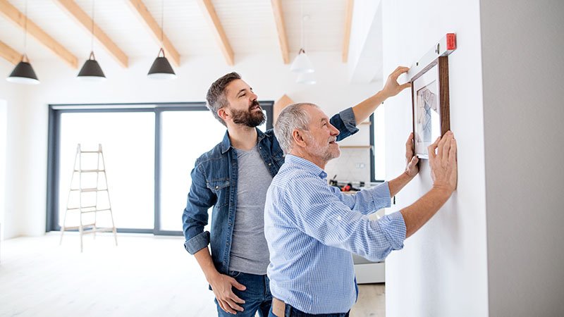 En far og hans voksne sønn henger opp et bilde på veggen i stua.