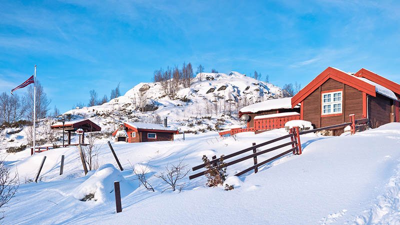 Snødekte røde hytter med fjell i bakgrunnen under blå himmel.