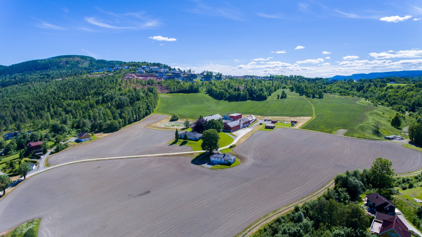 Dronebilde av stort gårsområde med jorder, grønne åkre, blå himmel og en gård midt i.