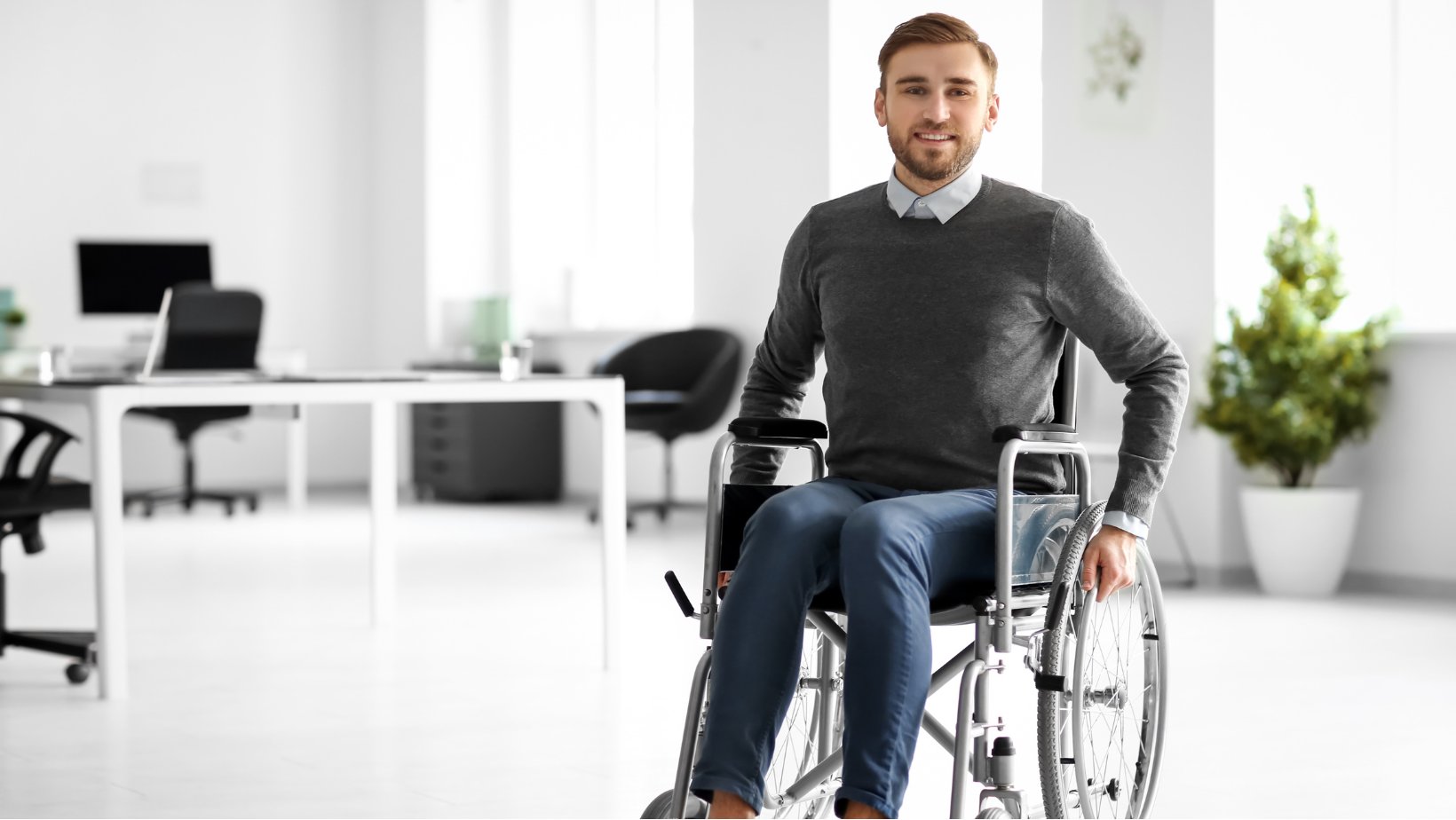 Person sitter i en rullestol i et lyst kontormiljø med skrivebord og stoler i bakgrunnen.