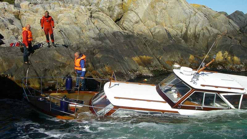 Motorbåt har gått på grunn ved steinete kyst, med tre personer i redningsvester og overlevelsesdrakter stående på land og ved båten.