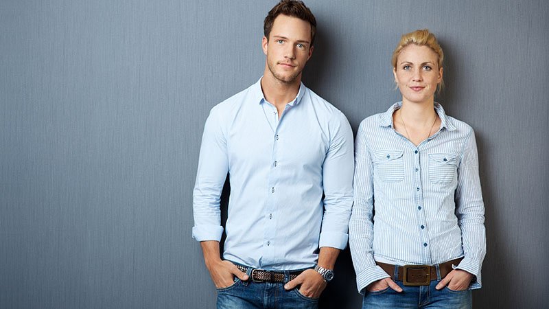 Mann og dame, begge i lyseblå skjorte og olabukser, står med hendene i lommene og ser i kamera.