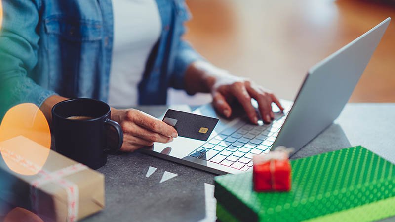 En mann bruker laptop og holder et kredittkort, med kaffekopp og gaver på bordet.