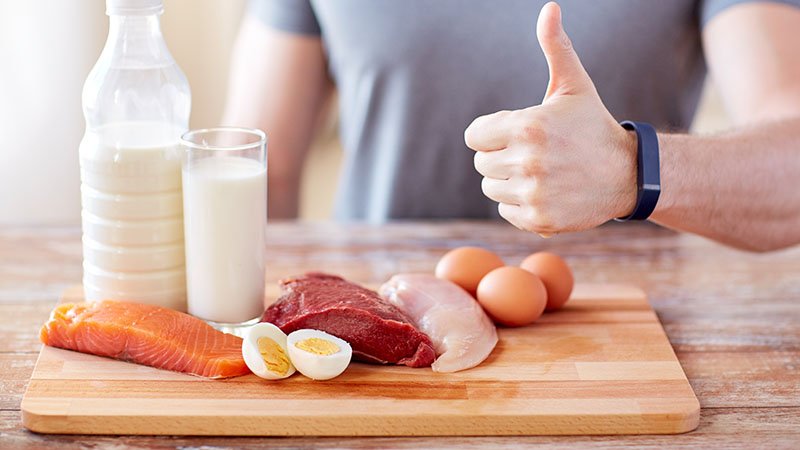 Hånd med tommel opp bak et skjærebrett med melk, egg, fisk og kjøtt.