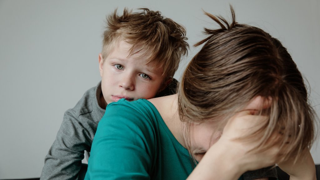 Gutt ser i kamera mens han lener seg på ryggen til moren, moren sitter med ansiktet i hendene