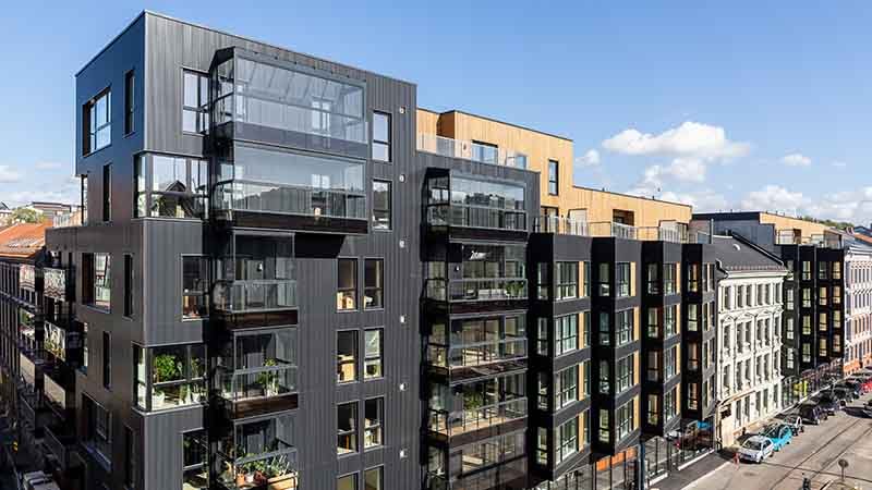 Moderne leilighetsbygg med mørke fasader og store glassvinduer, kombinert med trepanel på øverste etasje. Bygget ligger i en urban gate med klassiske bygårder på siden, og flere balkonger med planter er synlige.