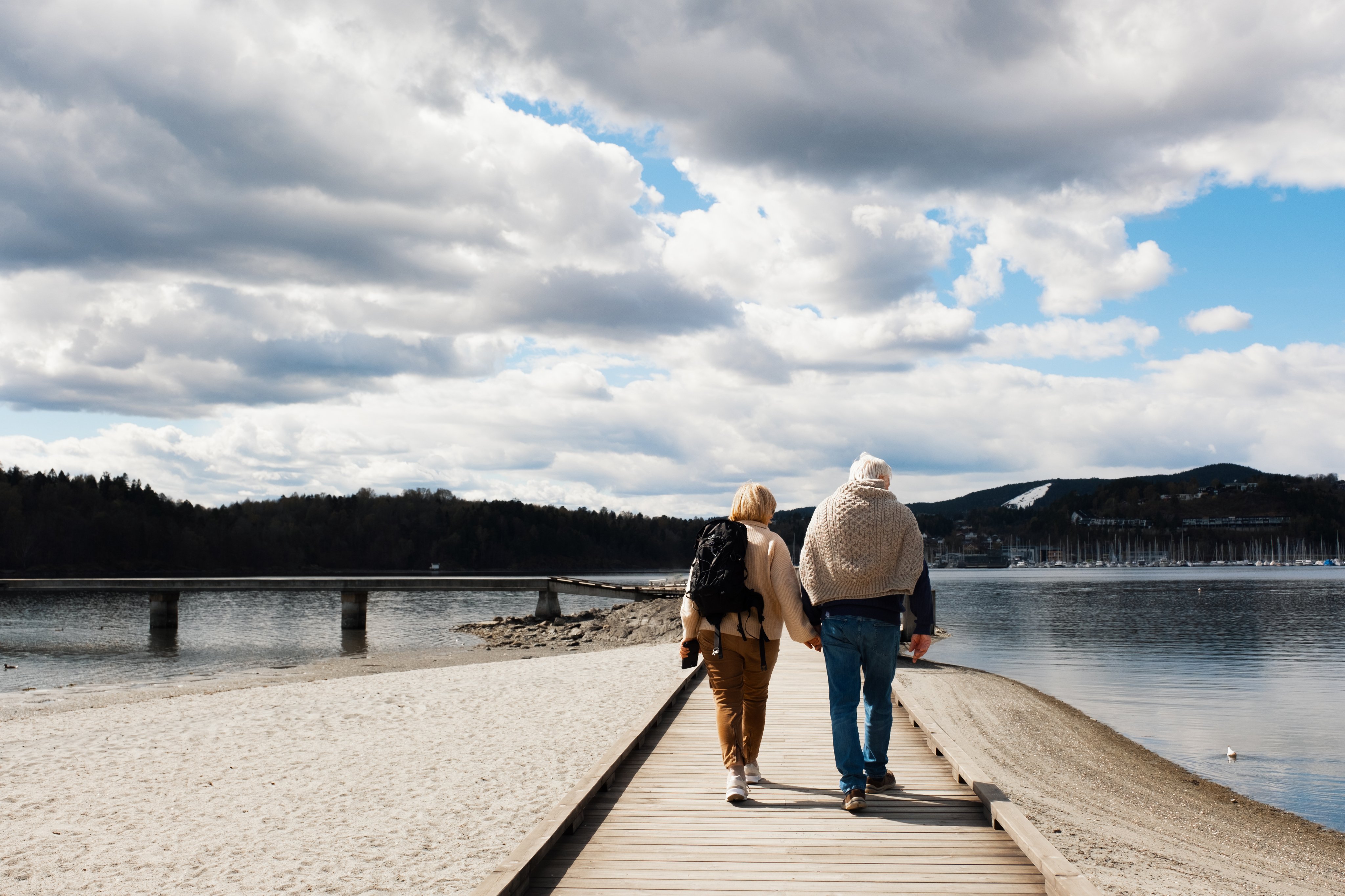 Et eldre par går langs en sjø, med båter og skog i bakgrunnen.