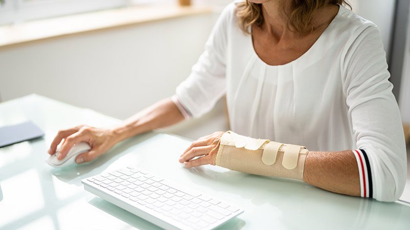 Ansatt med håndledsstøtte/bandasje på armen sitter ved skrivebord, og jobber med tastatur og mus.