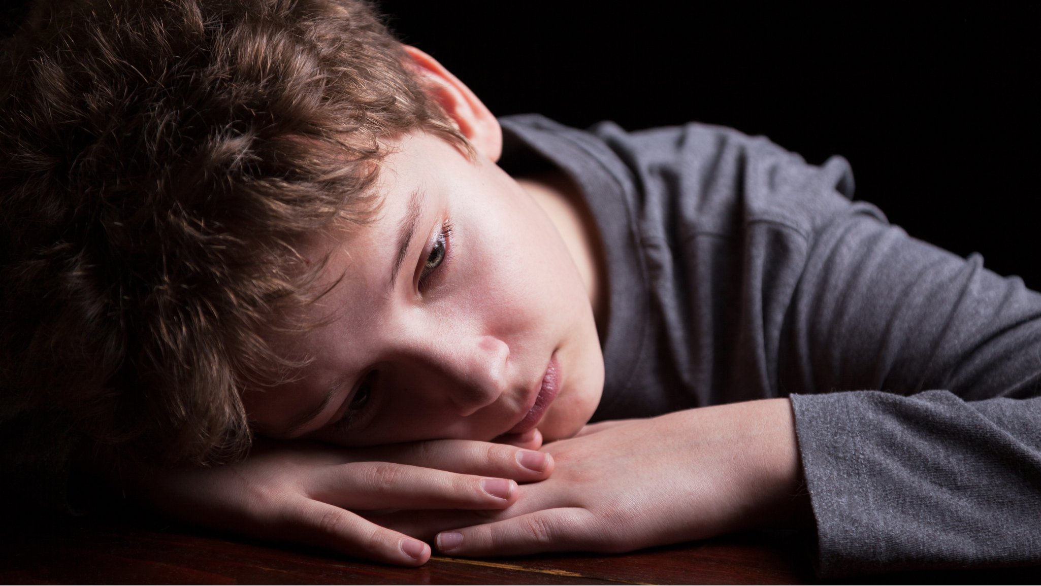 Ung tenåringsgutt lener hodet på hendene sine på et bord og ser trist ut.