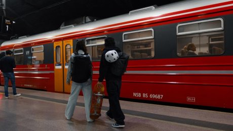 To personer står på en togplattform foran et NSB-tog, den ene holder et skateboard og har en hjelm festet til sekken