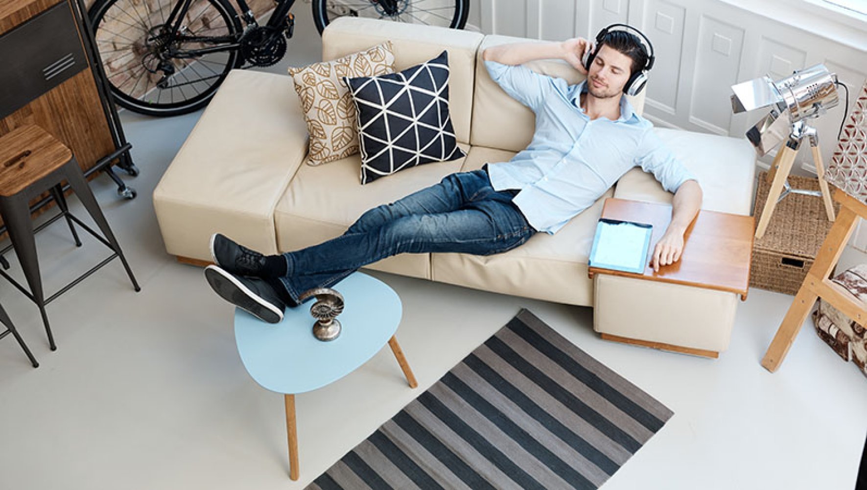 En ung mann ligger tilbakelent og avslappet i en sofa. Han har headset på hodet og bena på bordet.