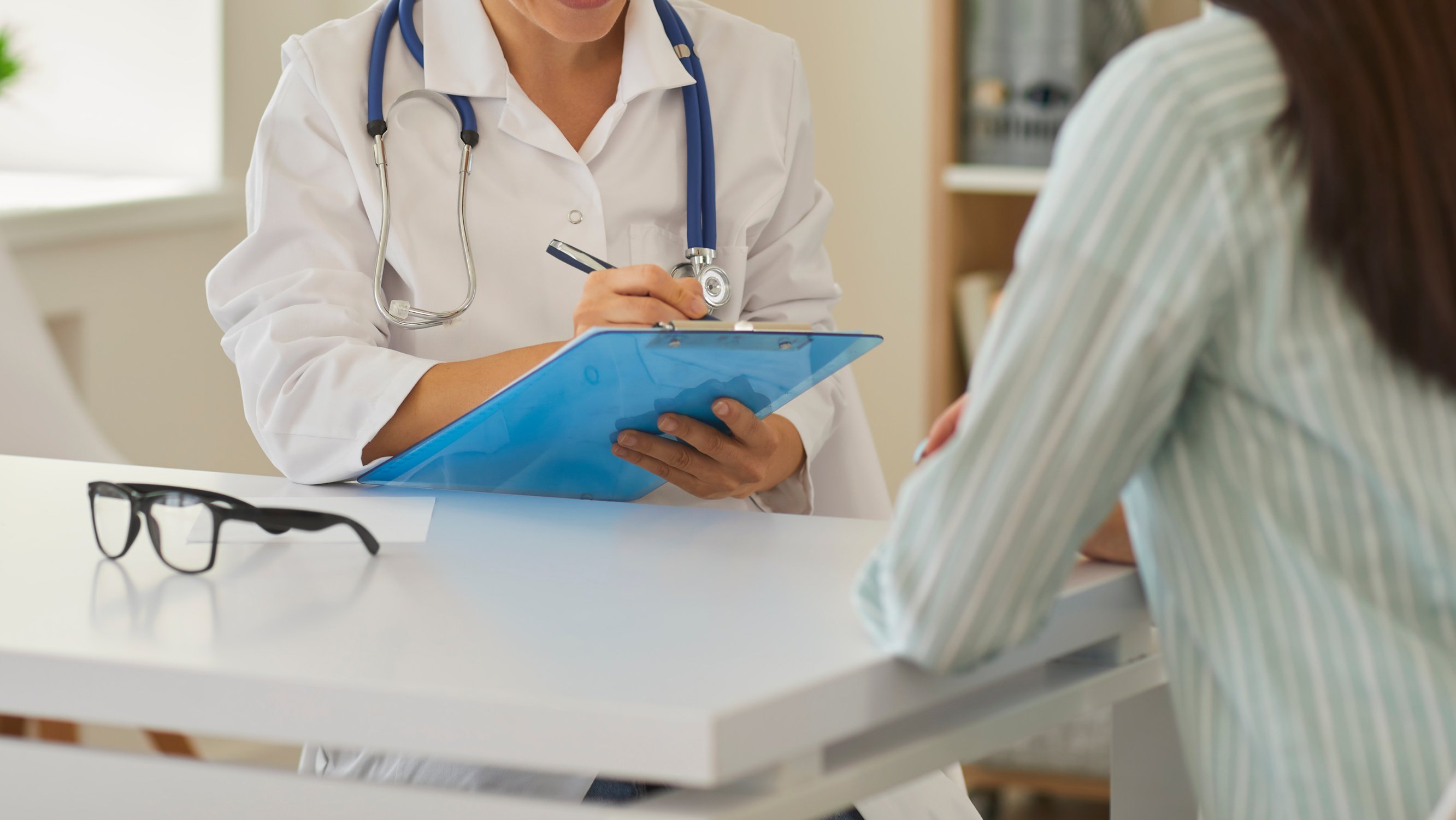 Lege med stetoskop skriver på et blått skjema under en konsultasjon.