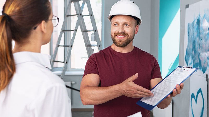 En person i rød t-skjorte og hvit vernehjelm holder en blå mappe med dokumenter og peker på innholdet, mens en annen person i hvit skjorte står foran. I bakgrunnen er en stige og en vegg under oppussing.
