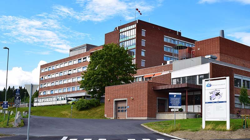 Stor, rød mursteinsbygning med flere etasjer, omgitt av grøntområde og skilt ved inngangen.