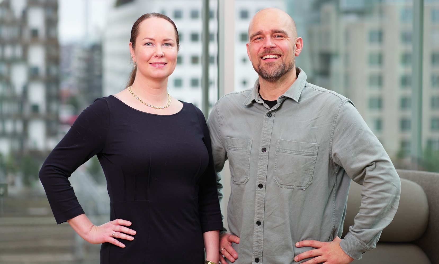 En mann og en kvinne står foran en stor glassvegg med urbane bygninger i bakgrunnen. Begge har hendene plassert på hoftene.