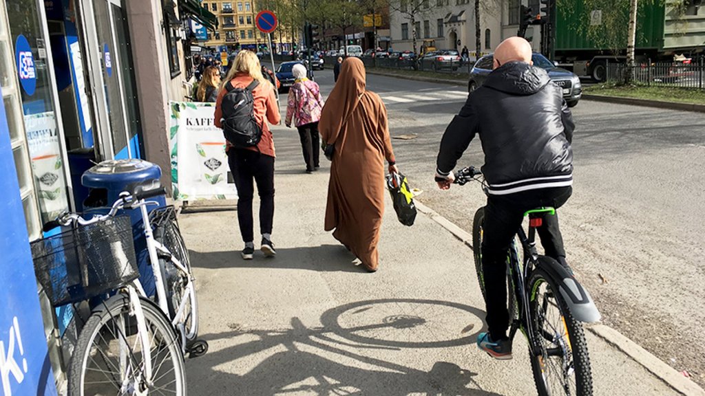 Folk går og sykler langs et fortau ved en butikk, med biler og bygninger i bakgrunnen.
