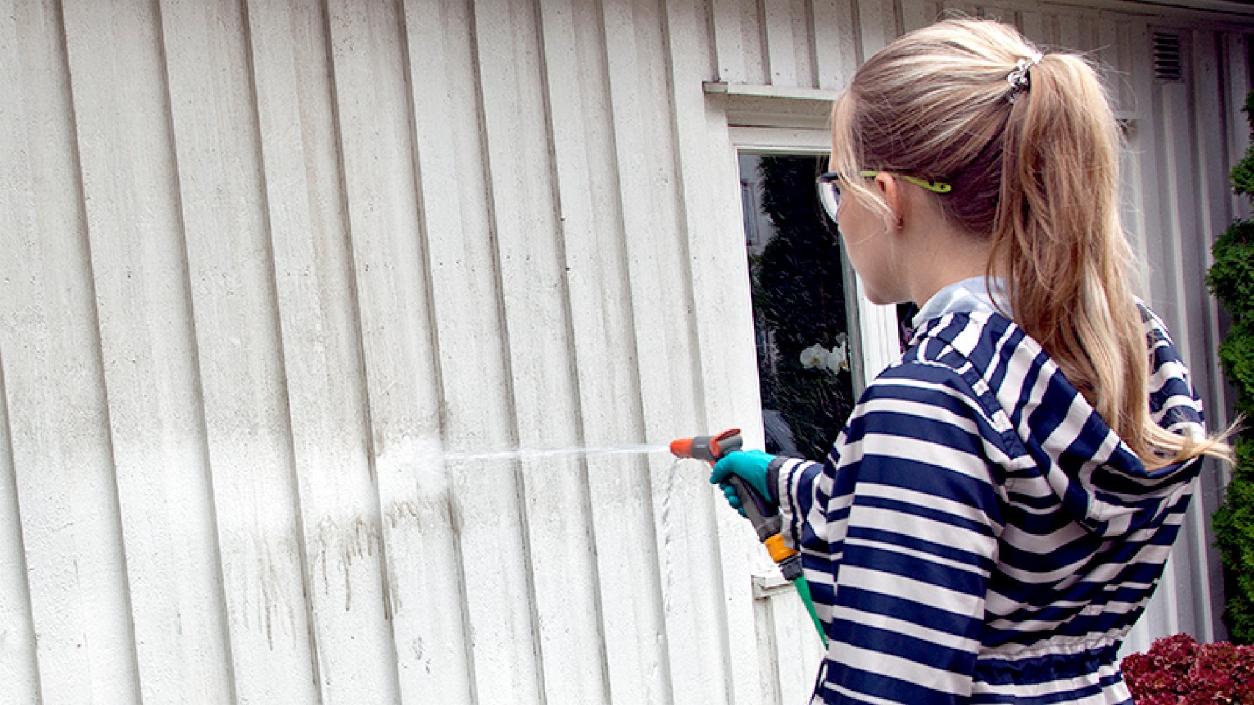 En kvinne spyler en skitten husvegg med hageslangen.