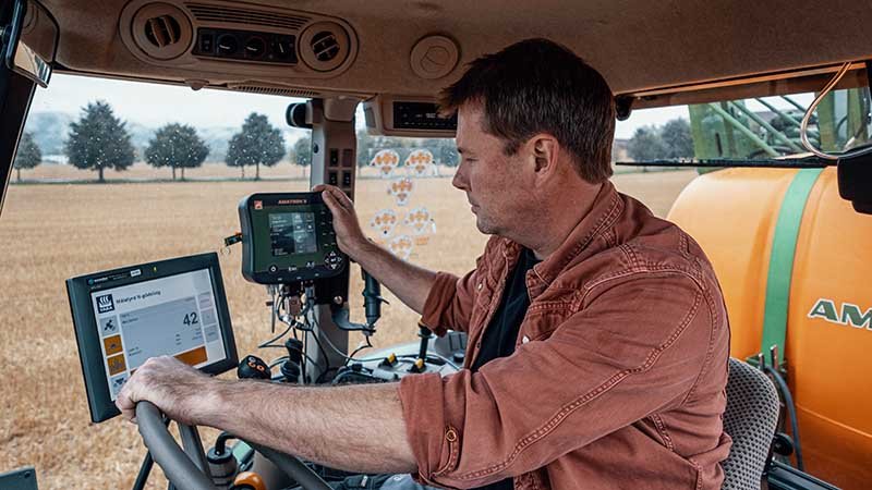 Person sitter i førerhuset på en traktor og betjener digitale skjermer, med åker og trær synlig gjennom vinduene.
