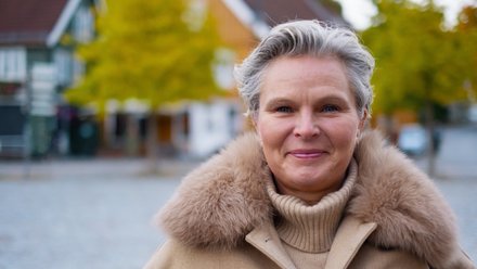 Person i brun kofte med pelskrage står ute med trær og bygninger bak.