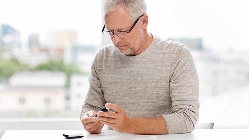 En mann sitter ved et bord og bruker et blodsukkermåler-sett med teststrimler og måleapparat.