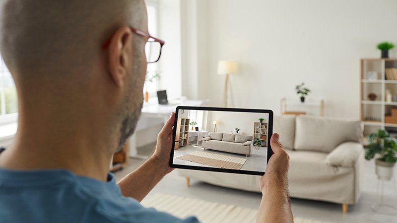 En mann holder et nettbrett som viser et bilde av stuen foran ham.