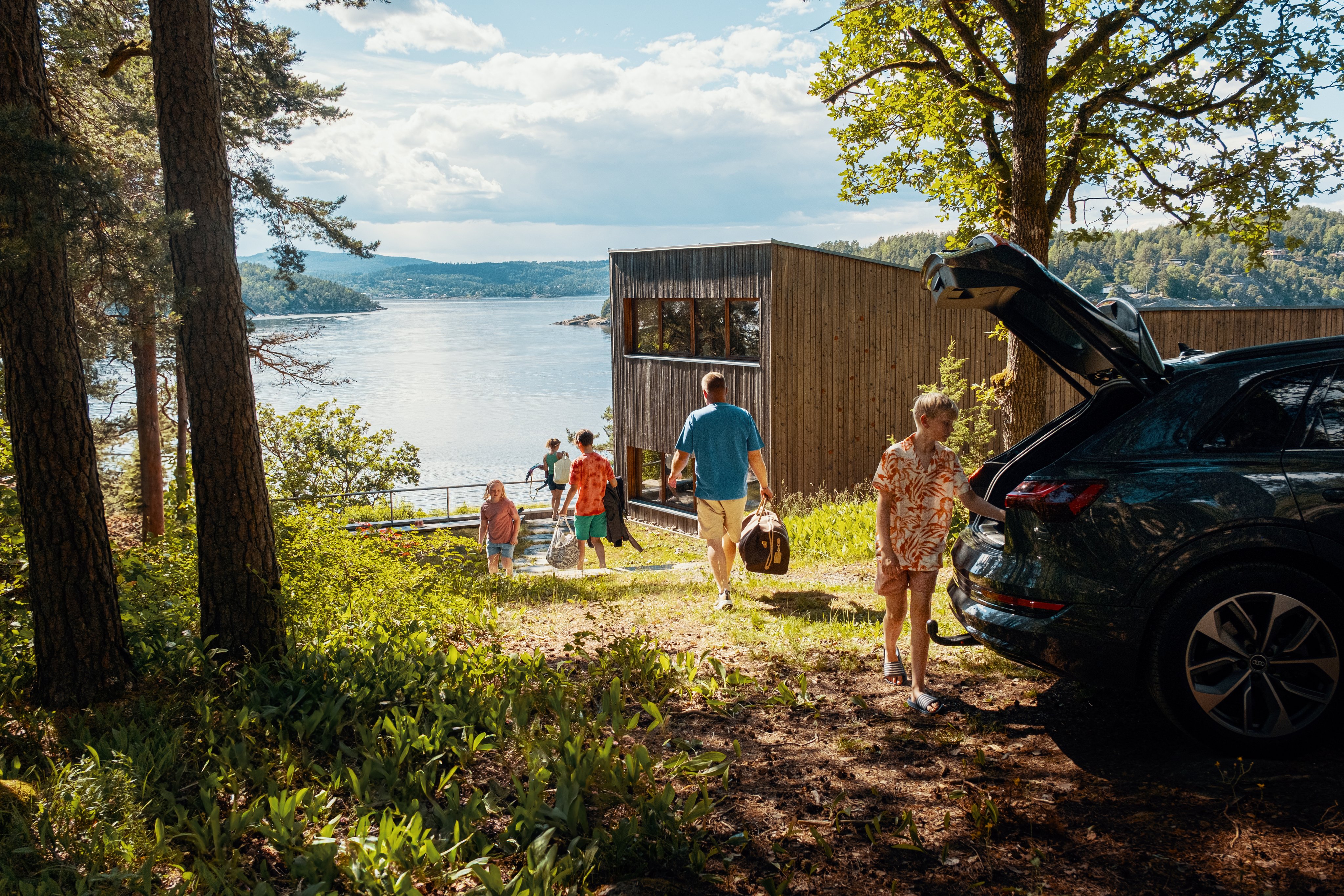 Familie ankommer en hytte ved vannet, med bil parkert i forgrunnen.