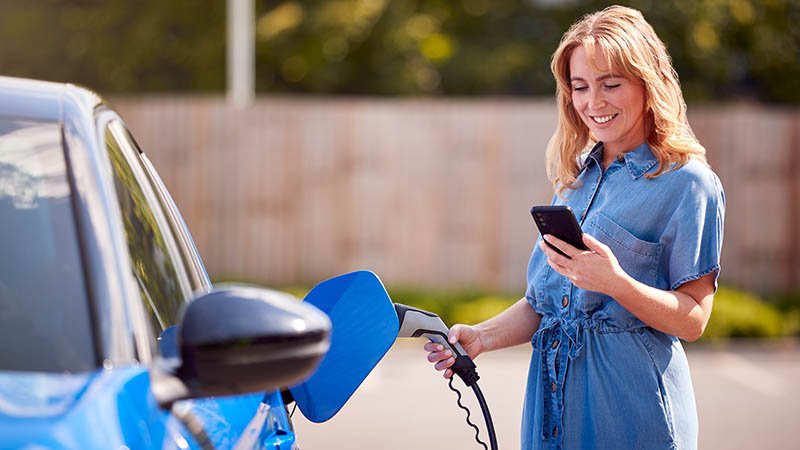 Smilende kvinne står ved siden av en elbil med ladeluka åpen, med en elbillader i en hånd og mobilen i den andre.