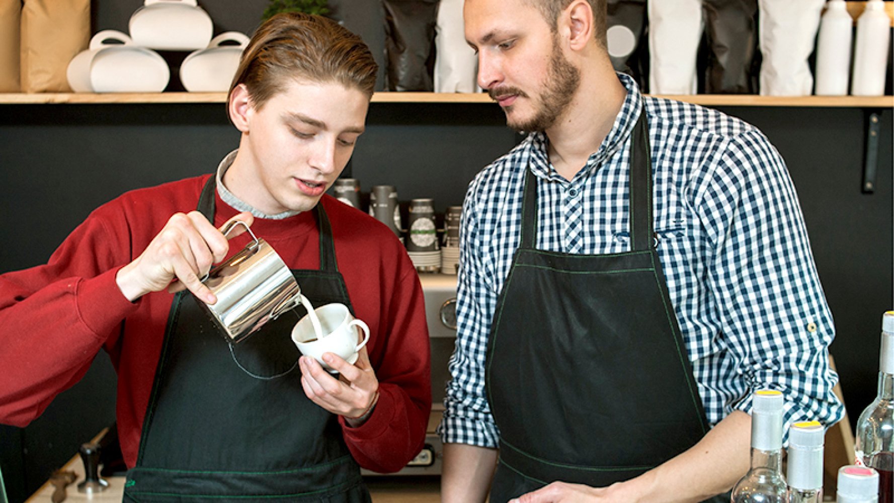 Ung barista i en kaffebar heller melk i en kaffekopp mens en eldre barista står ved siden av og ser på