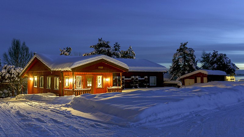 En snødekt fjellhytte i skumring hvor lyset er slått på inne.