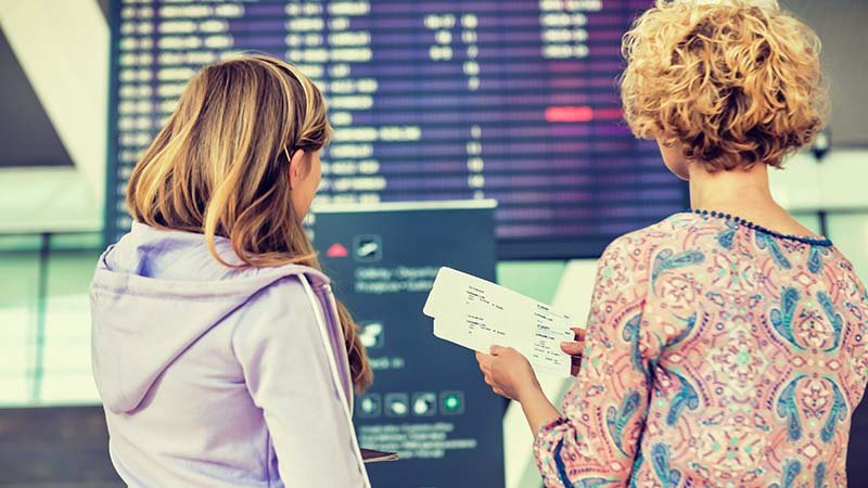 Mor og datter med ryggen til kamera står med to flybilletter i hånden og ser opp på en tidstabell på flyplassen