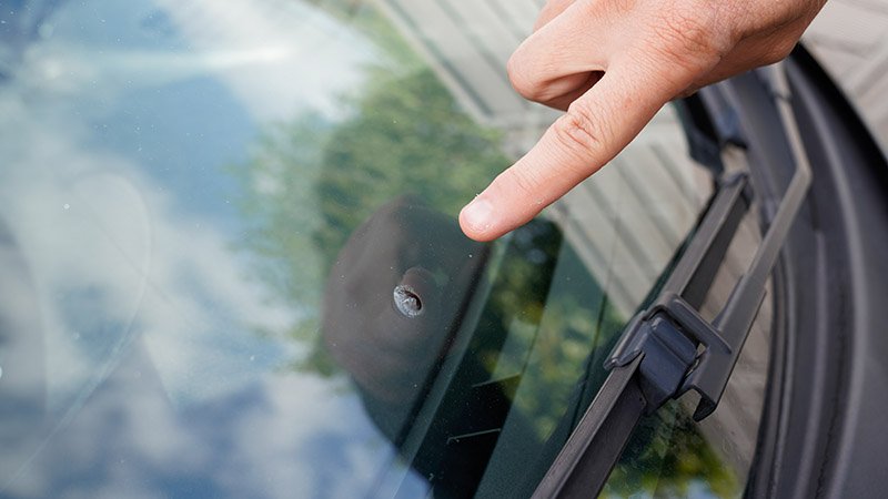 Finger peker på en steinsprutskade på frontruten på en bil.