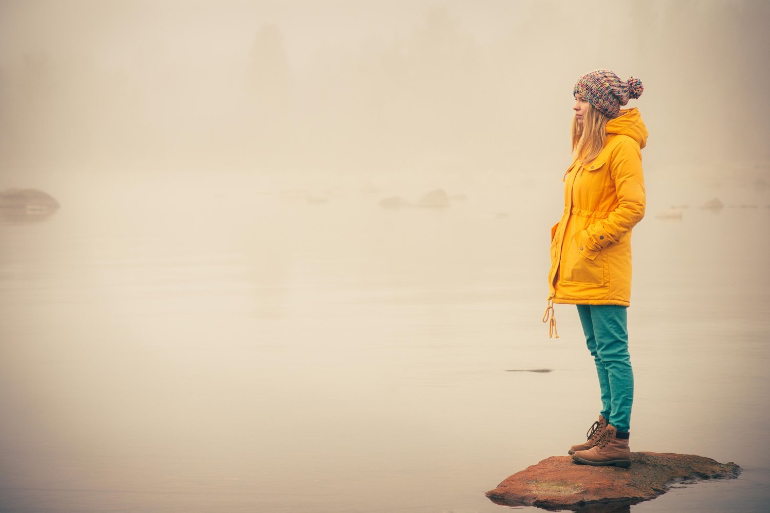 Kvinne i gul jakke stående på en stor stein ved havet